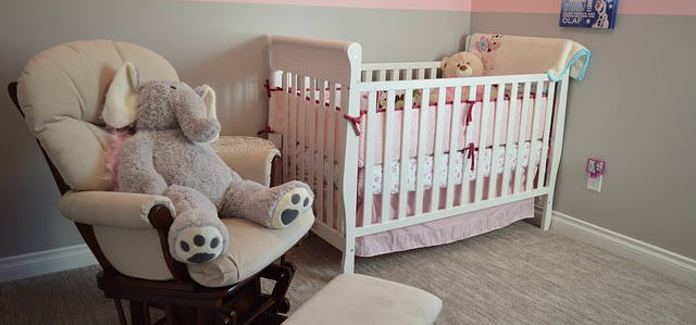 Ein Sessel und ein babybett in einem Raum. Auf dem Sessel sitzt ein Teddy. Das Babybett sind weiblich aus., wegen den Farben rosa und weiß.