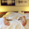 Füße schauen aus einem Bett heraus. Wenn sie gehören weiß man nicht. Vermutlich schläft die Person. Sie liegt auf dem Bauch, da die Füße nach unten zeigen.