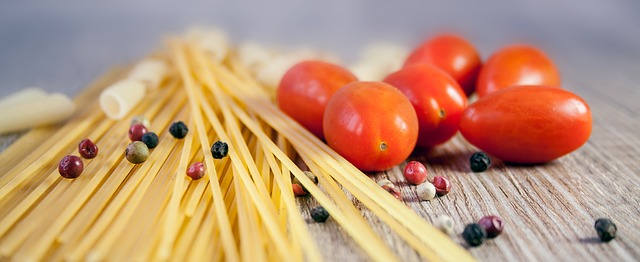 Spaghetti liegen auf einem Hölzernen untergrund. Sie sind ausgepackt . Daneben liegen kleine Tomaten. Verstreut sind Pfefferkörner zu finden.