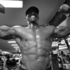 Ein schwarz weiß Bild. Ein Mann spannt seine Muskeln an und präsentiert sie im Fitnessstudio. Dabei hat er die Arme gehoben um seinen recht beeindruckenden Bizeps zu zeigen.