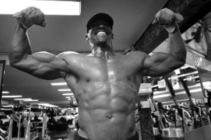 Ein schwarz weiß Bild. Ein Mann spannt seine Muskeln an und präsentiert sie im Fitnessstudio. Dabei hat er die Arme gehoben um seinen recht beeindruckenden Bizeps zu zeigen.