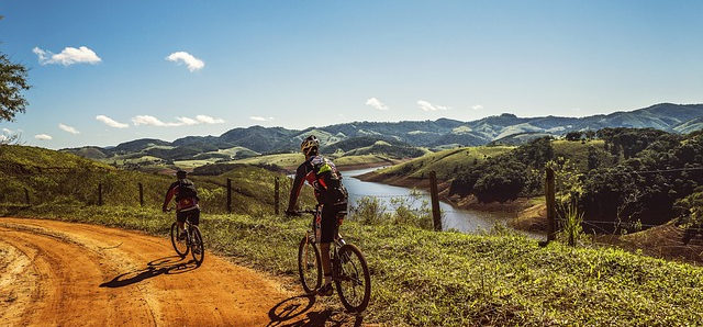 Zwei Radfahrer fahren durch eine Landschaft. Grüne Hügel und ein Fluss sind zu sehen. Der Weg auf dem die Radfahrer fahren ist sandig und steinig.