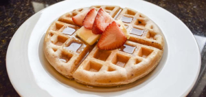 Eine Waffel auf einem Teller. Beide haben eine runde Form. Auf der Waffel ist Sirup und eine Erdbeere.